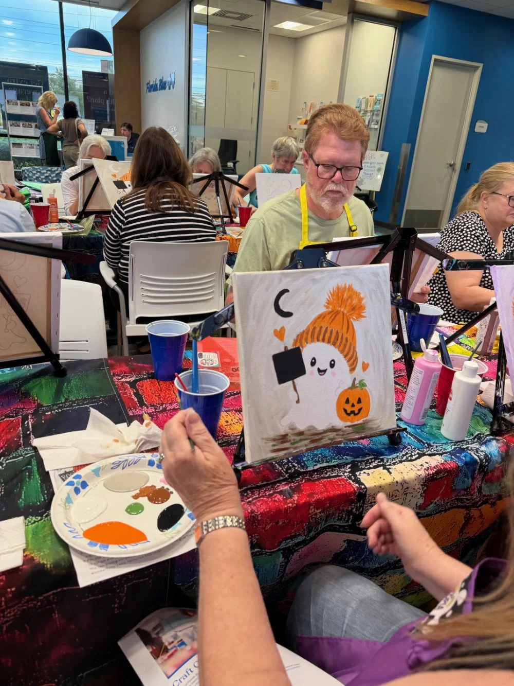 People participating in a paint and sip event, painting Halloween-themed pictures, with a focus on a painting of a cute ghost with a pumpkin and a hat, on a colorful table with art supplies.