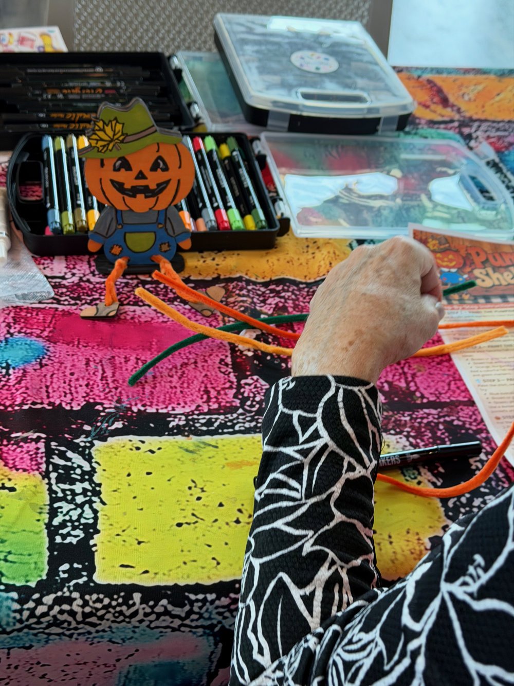 Colorful craft table with markers, paper, and a Halloween-themed paper decoration of a pumpkin character dressed as a scarecrow. A person is holding orange pipe cleaners, and there are plastic containers with craft supplies in the background.