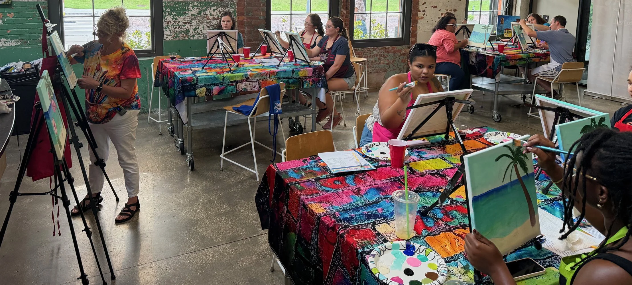 People participating in a painting class, creating artwork on canvases at tables covered with colorful, artistic tablecloths.