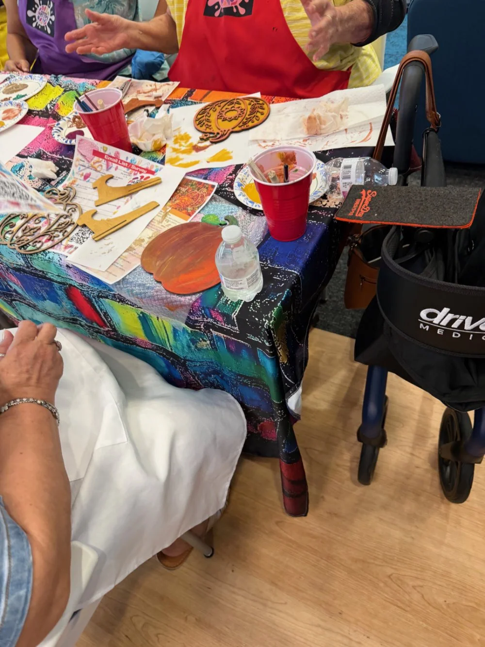 Decorative table with arts and crafts supplies, paper plates, drinking cups, water bottle, and a wheelchair nearby, likely for a crafting activity.