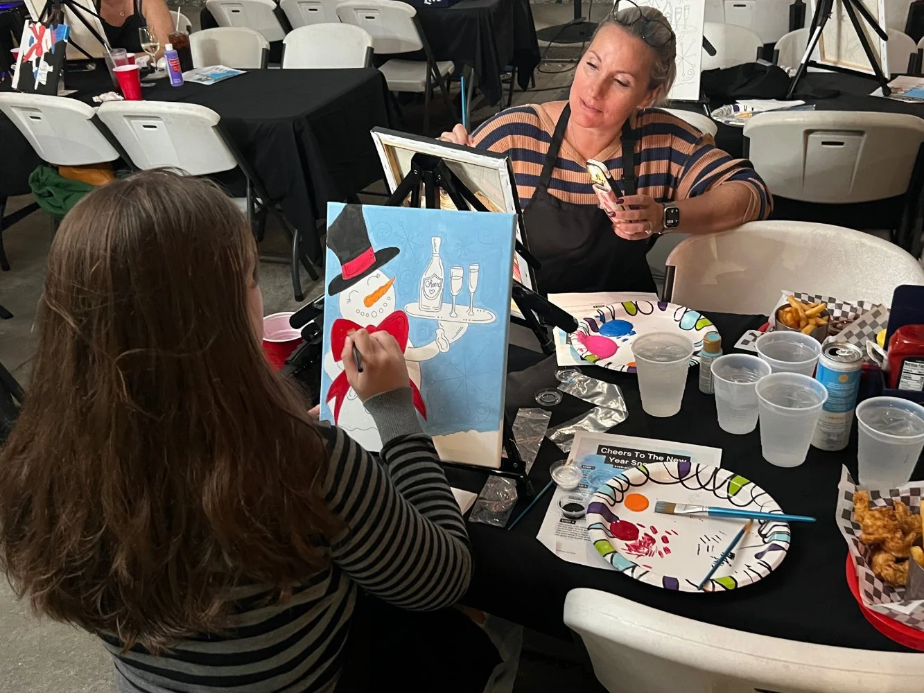 A girl painting a snowman wearing a black top hat, red scarf, and orange nose, while a woman with a striped shirt and apron looks on at a table decorated with snacks and drinks, in an art event setting.
