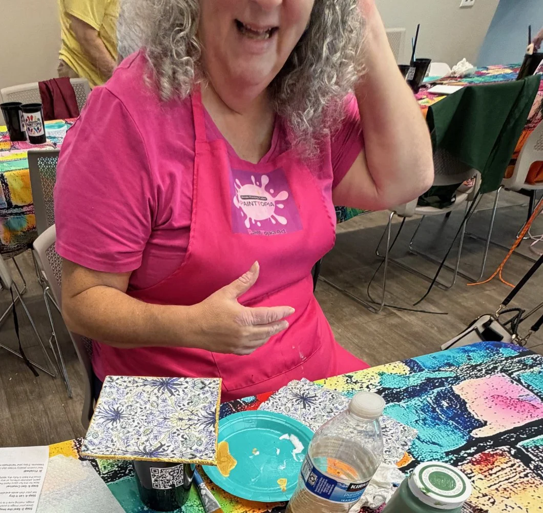 Woman sitting at a table wearing a pink apron with a paint splash design; table covered with colorful painted fabric, paint supplies, and a small plate; woman smiling and making a gesture with her hand; background shows chairs and another person in a