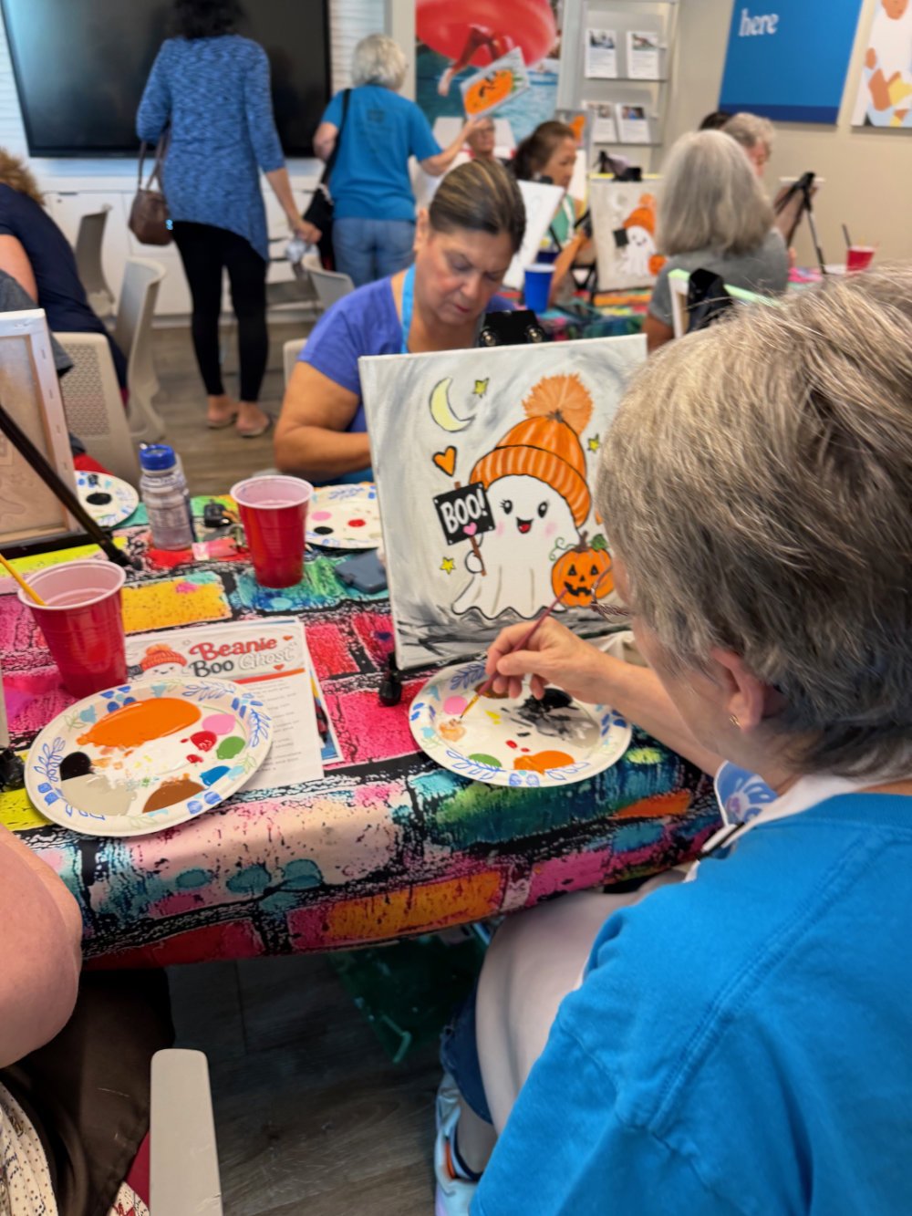 People participating in a Halloween-themed paint-and-sip event. They are painting spooky pumpkin and ghost images. The table has Halloween decorations, paint palettes, brushes, and red cups.