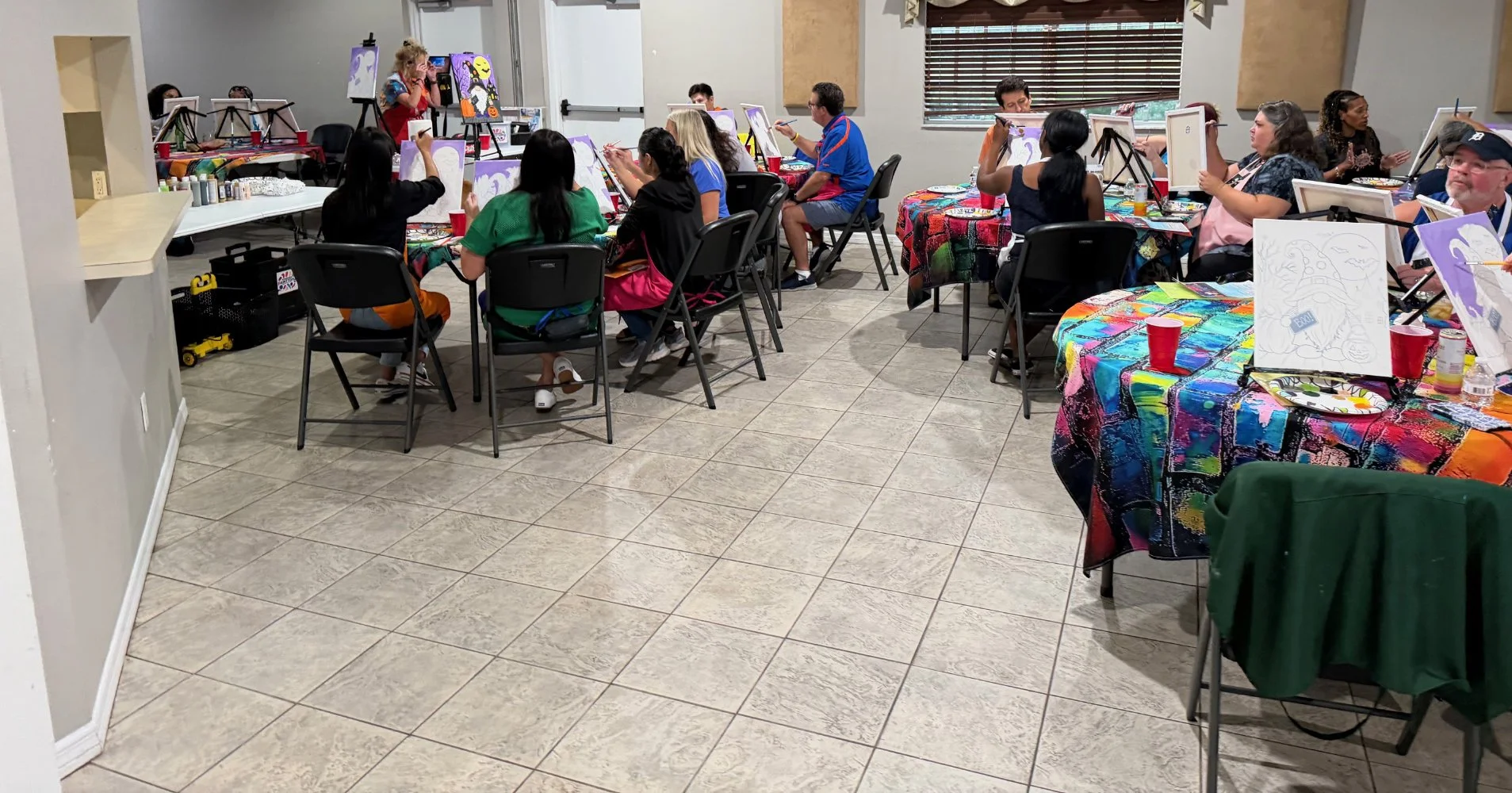 People participating in a group painting class in a room with colorful tablecloths, easels, and completed artwork, suggested as a lively and creative event.