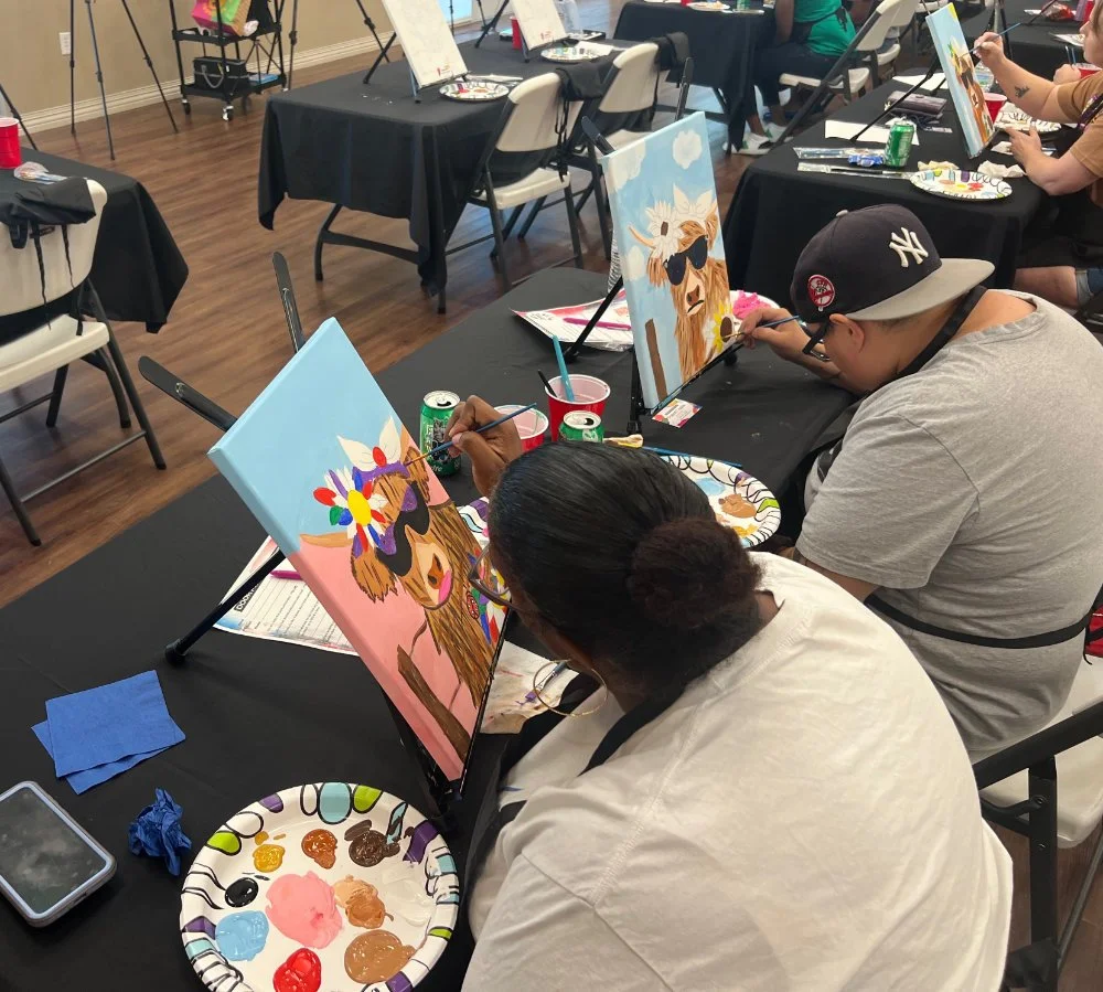 People painting animal portraits at a group art class, with canvases featuring a lion wearing sunglasses and a colorful headdress, set on a black table with paint palettes, cups, and art supplies.