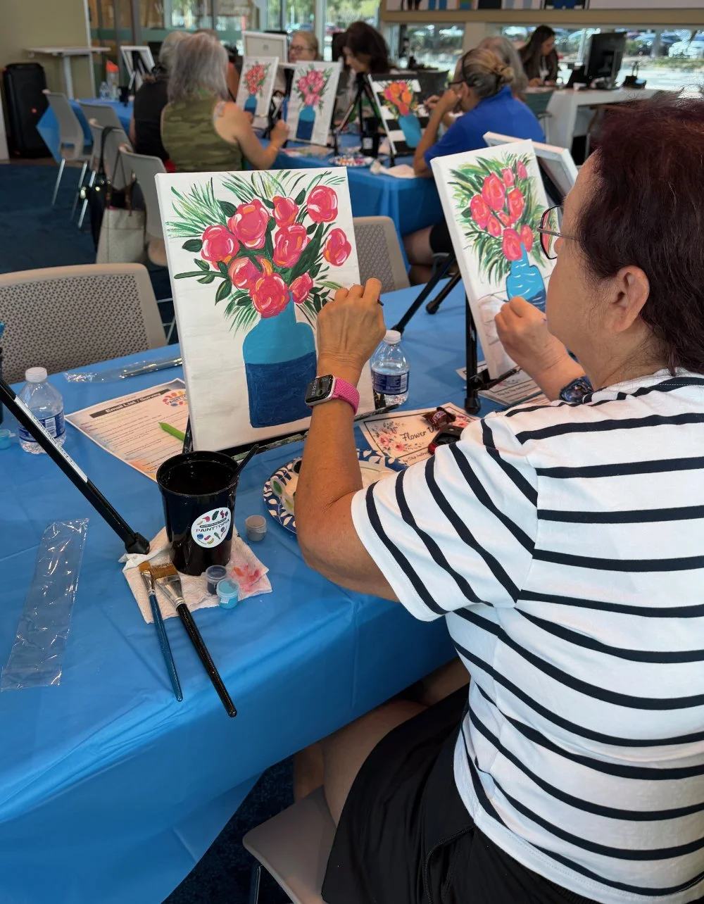 Woman in a striped shirt painting a colorful floral still life on canvas at a group art class, with other participants working on similar paintings in the background.