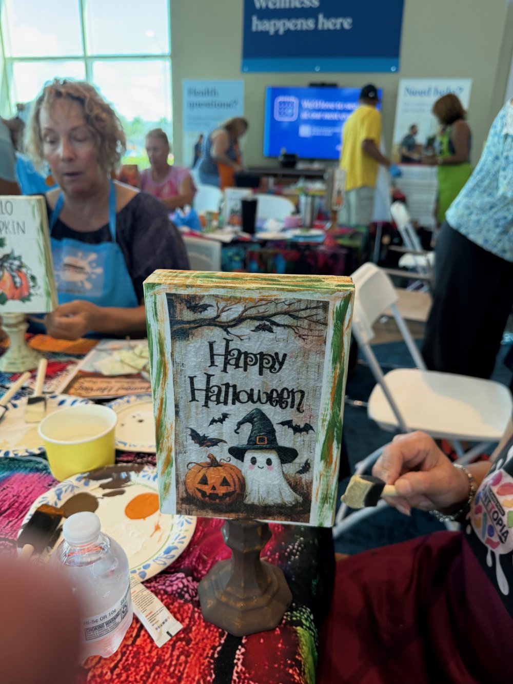 A Halloween-themed sign with the message "Happy Halloween" featuring a witch hat-wearing ghost, a carved pumpkin, and flying bats on a table at a craft event.