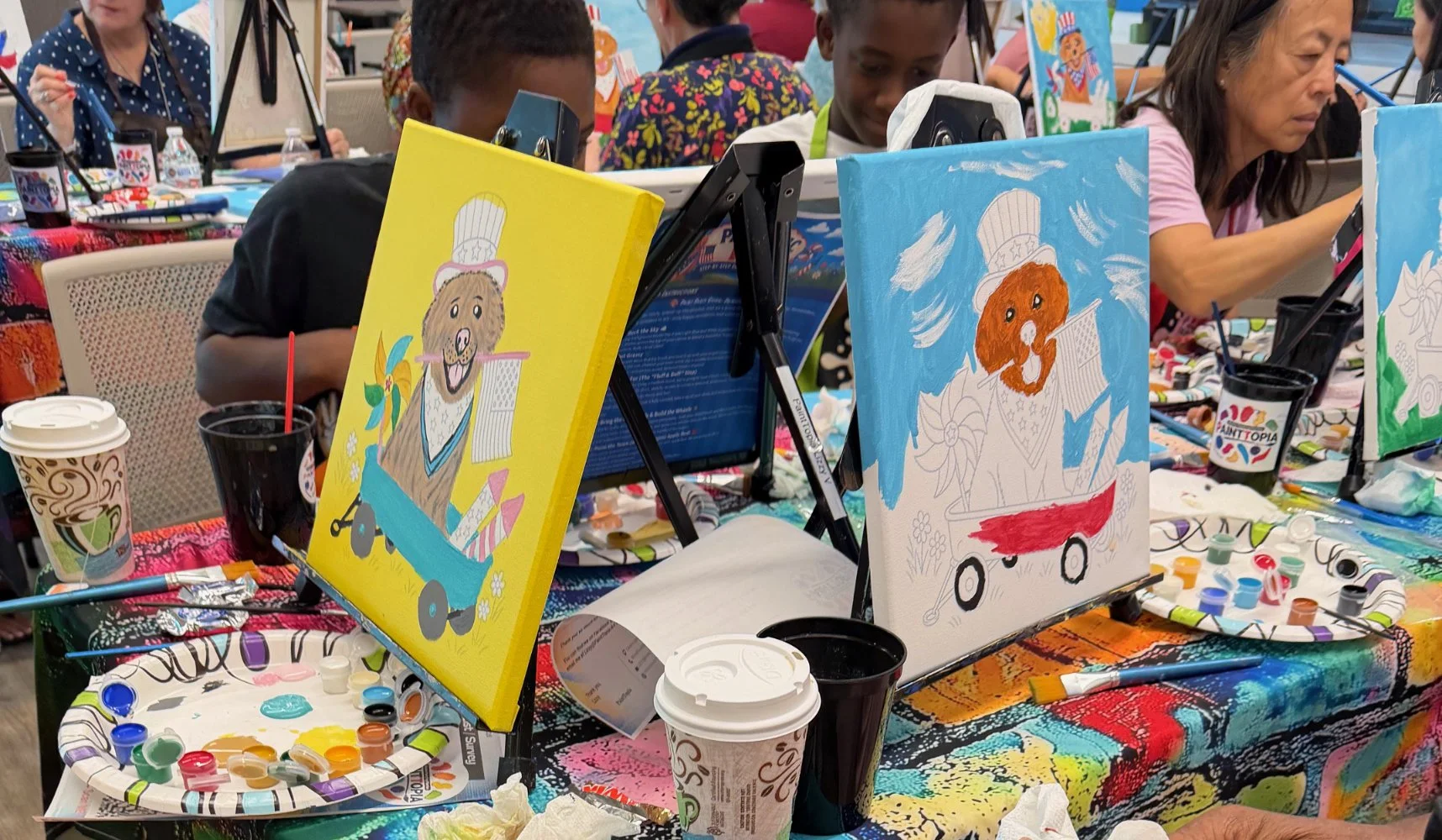 Children and adults participating in a painting activity at an art event, with colorful paintings of dogs in wagons displayed on small easels on a table covered with a vibrant, multicolored tablecloth, surrounded by art supplies, paper coffee cups, a