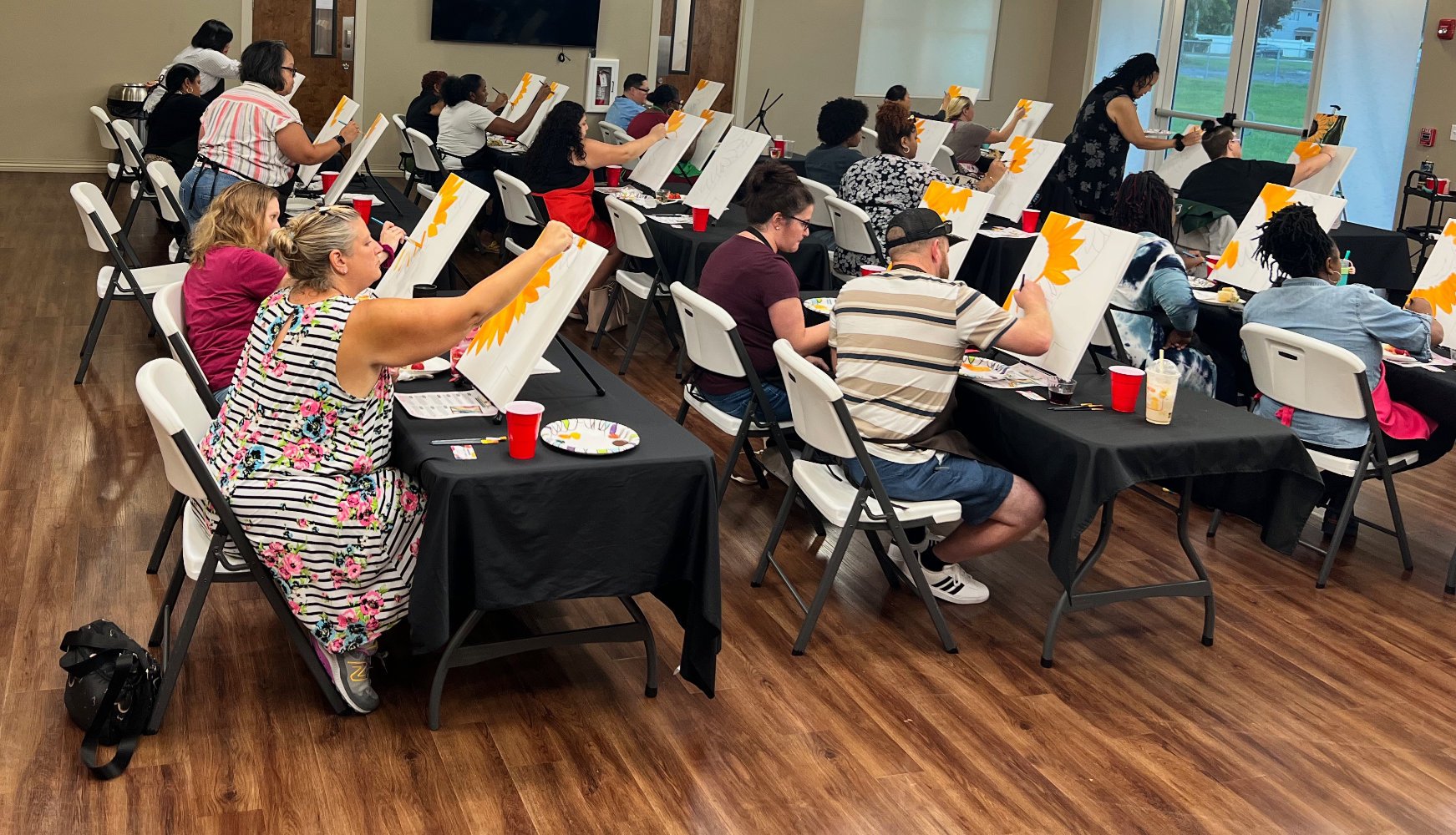 People painting on canvases set up on easels indoors, with some viewers capturing their work with cameras, during a group painting event.