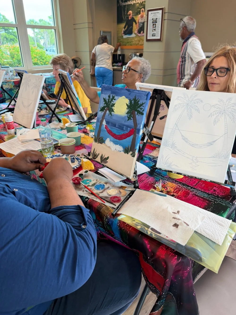Group of people participating in a painting class at a table, with paintings of a beach scene, a sunset, and a hammock on display. The participants are focused on their canvases, and painting supplies are scattered on the table.