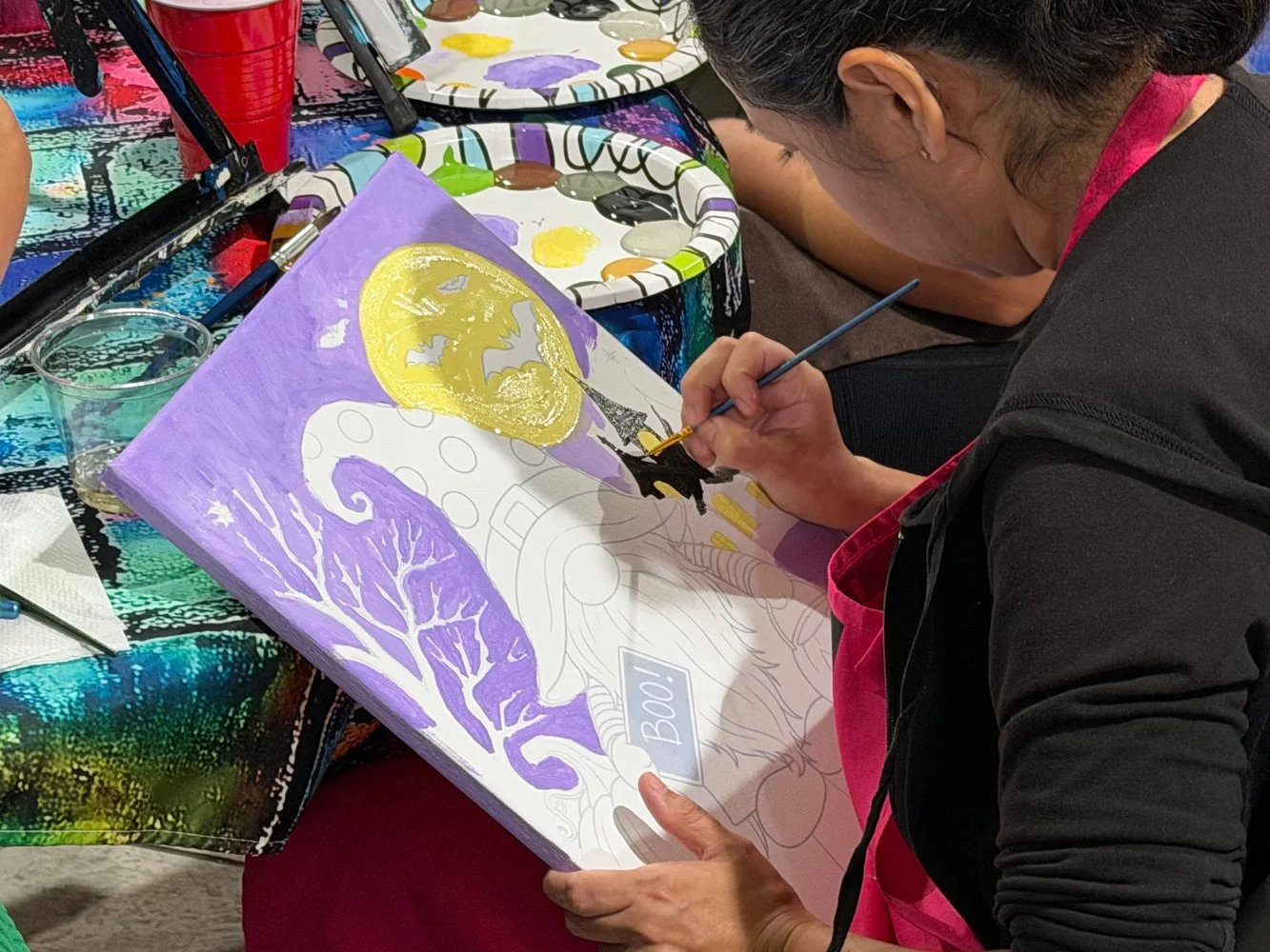 A person is painting a Halloween-themed picture on a canvas, featuring a large yellow moon, black bat silhouettes, a black cat, a witch's hat, and a gnarled tree; the artist is in the process of adding details.