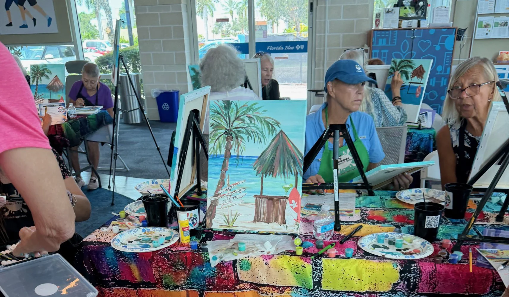 Group of people painting tropical beach scene at an art class with easels, canvases, and art supplies inside a room with large windows and viewed parking lot outside.