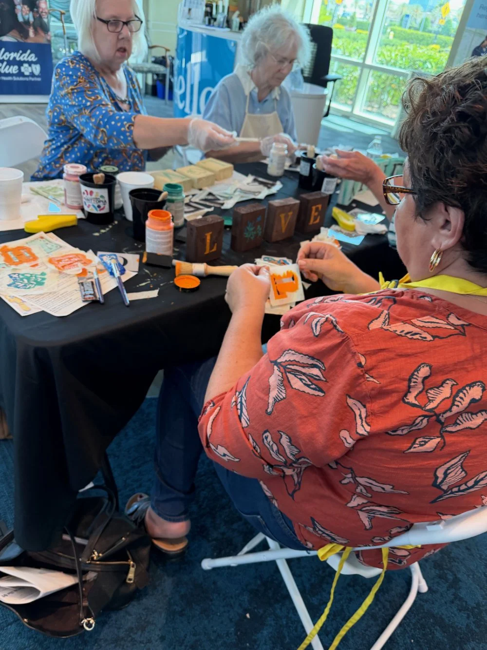 Group of women crafting at a table, decorated with painted wooden blocks spelling 'LOVE', various craft supplies, and art projects, in a bright room with large windows.