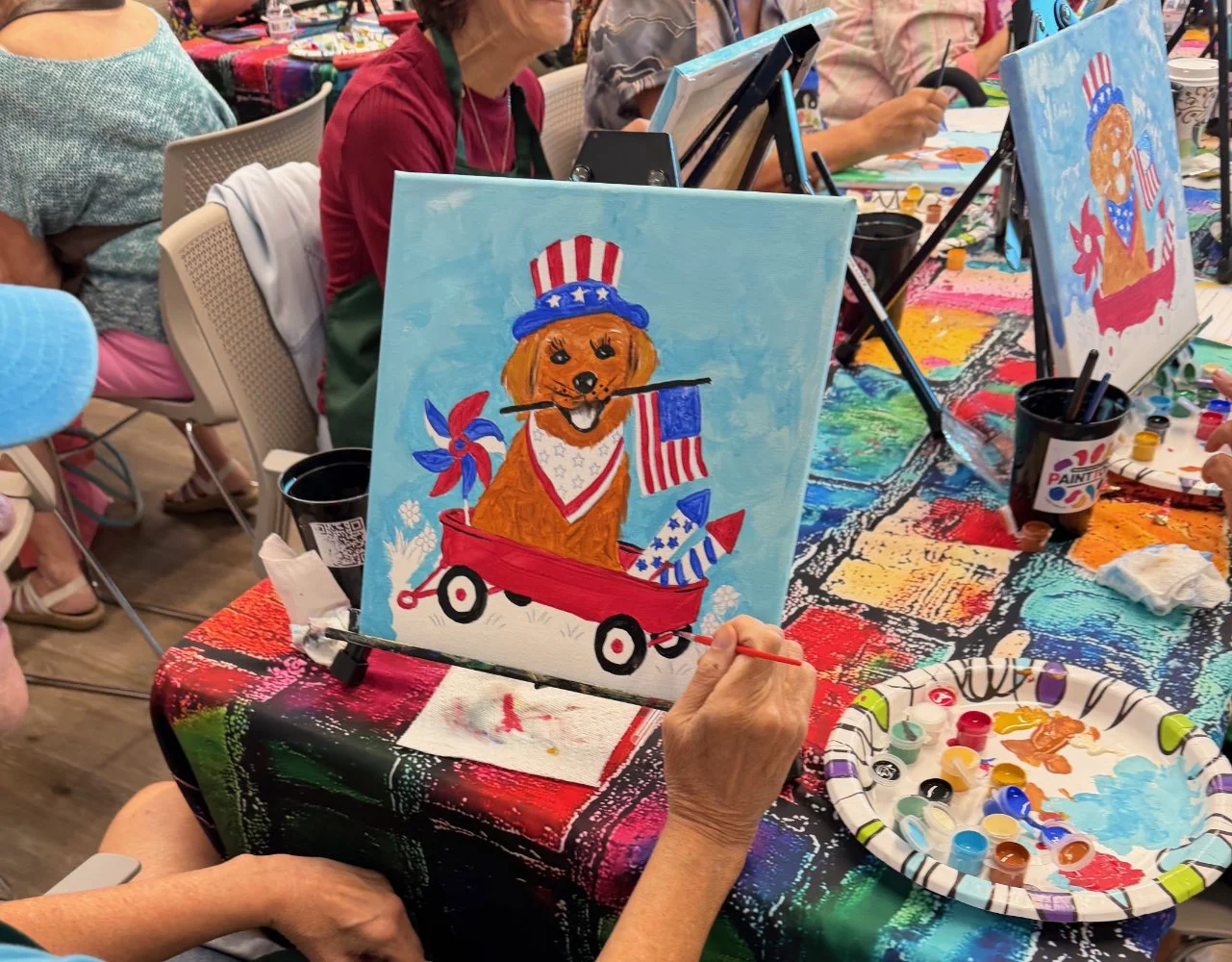 A woman is painting a picture of a dog dressed in patriotic clothing, sitting in a red wagon with American flags and a pinwheel, at a group art event. The table has paint supplies and a colorful tablecloth.