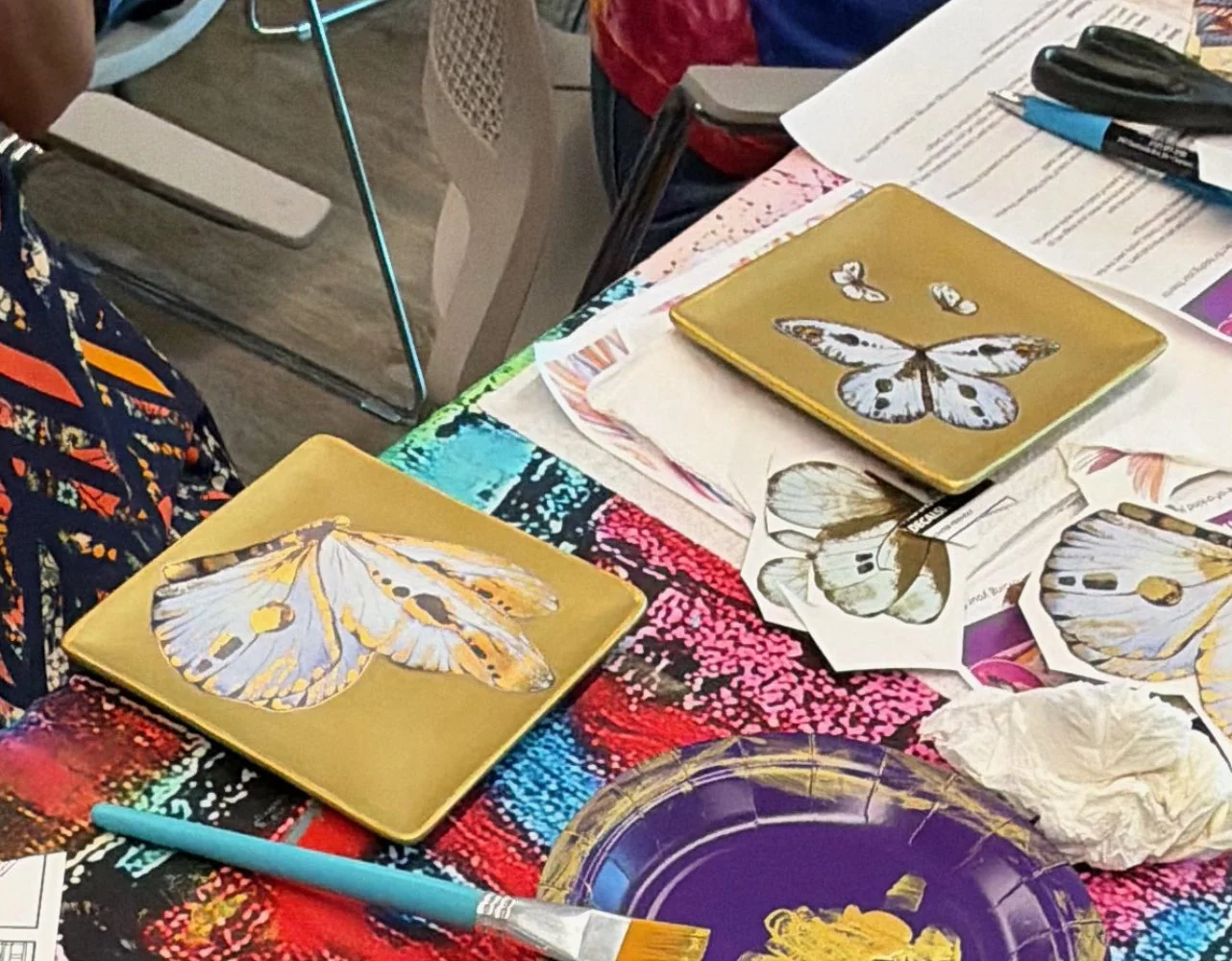 Table with butterfly-themed art supplies, including two gold-colored plates with butterfly illustrations, scattered cutouts of butterflies, paintbrush, and papers.