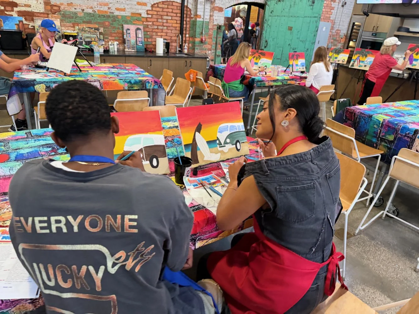 People participating in a painting class, with supplies and several colorful landscape paintings, in an industrial-style room with brick walls and wooden accents.