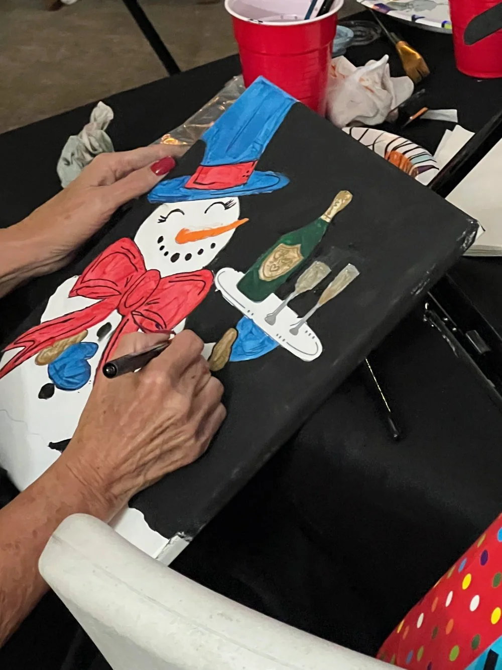 A person with red painted nails is painting a festive snowman decoration on a black canvas. The snowman has a top hat, a red bow, and an orange carrot nose, with a background that suggests a holiday or winter scene.