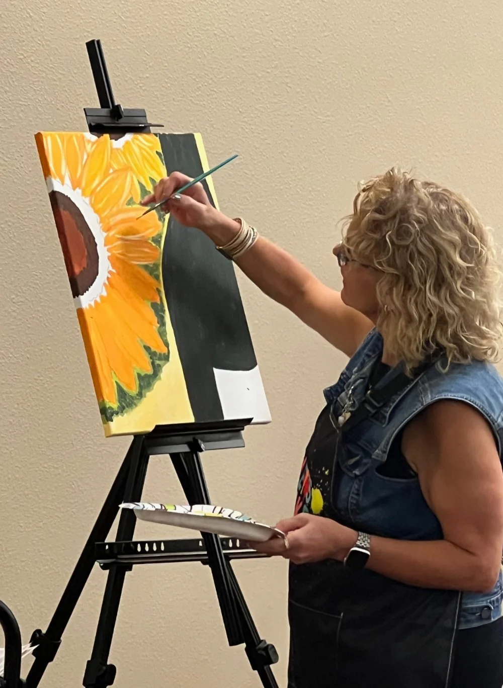 A woman with curly blonde hair painting a sunflower on a canvas, standing in front of a beige wall, holding a palette in one hand and a brush in the other.