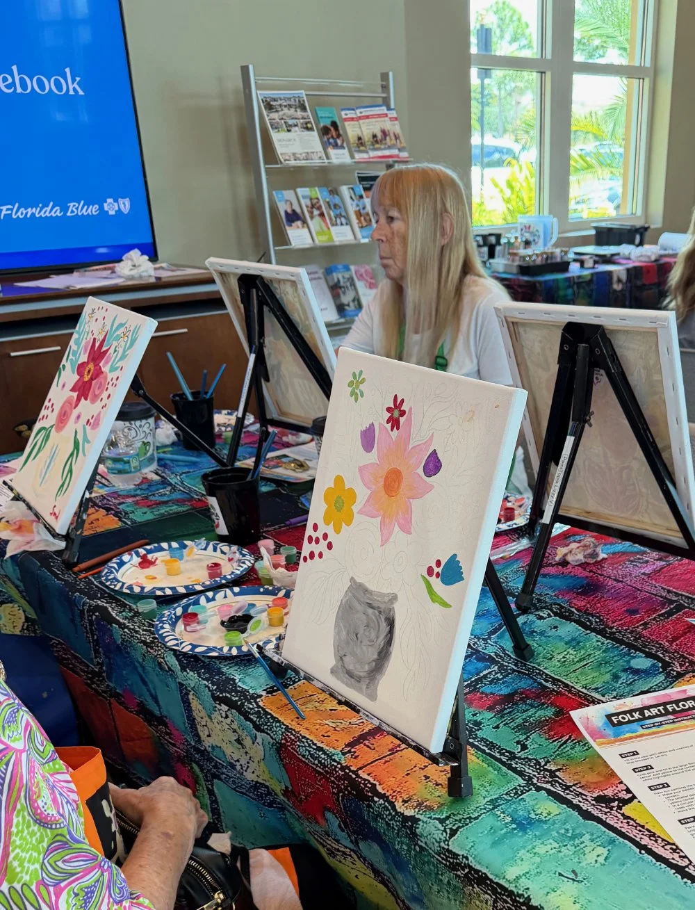 People participating in a folk art painting class with colorful floral paintings on canvases, paint palettes, and brushes on tables covered with vibrant multicolored tablecloths, inside a bright room with large windows.