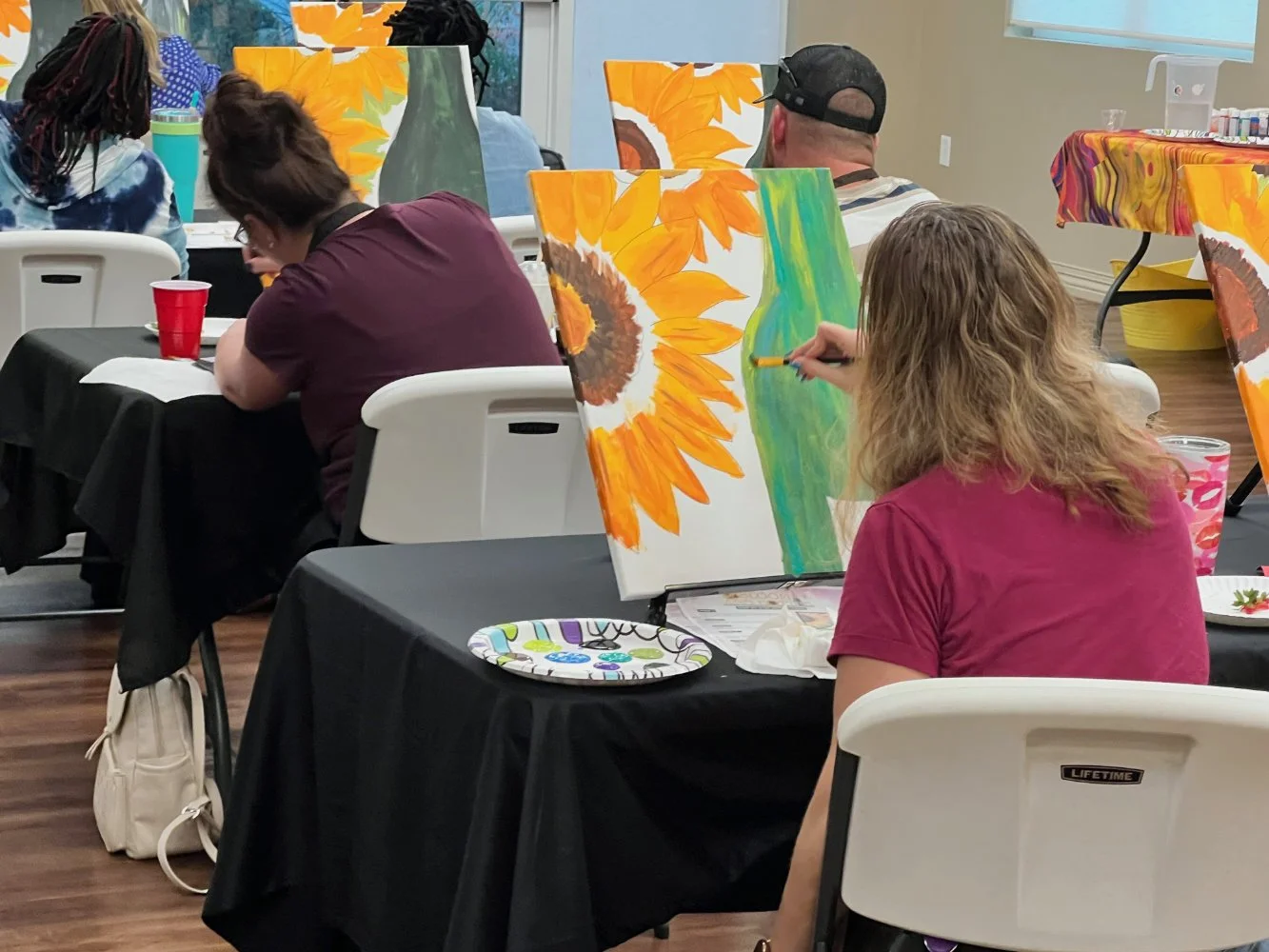People participating in a painting class, creating sunflower paintings on canvases with bright colors, sitting at black-covered tables in a well-lit room.