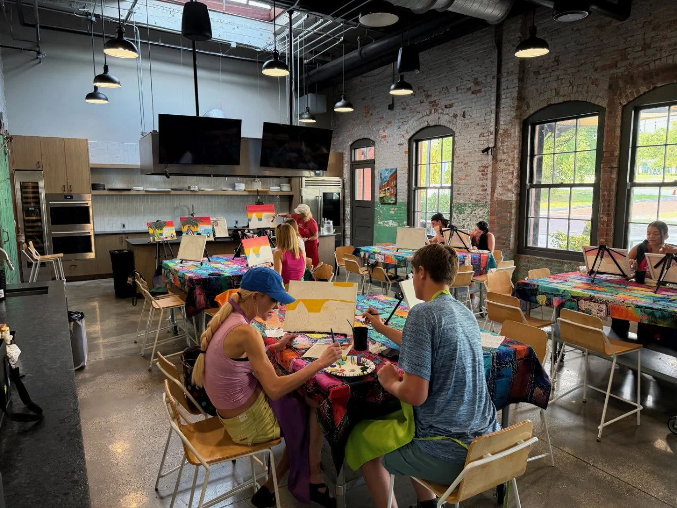 People painting at tables during a painting class inside an industrial-style studio with brick walls, large windows, and colorful tablecloths.