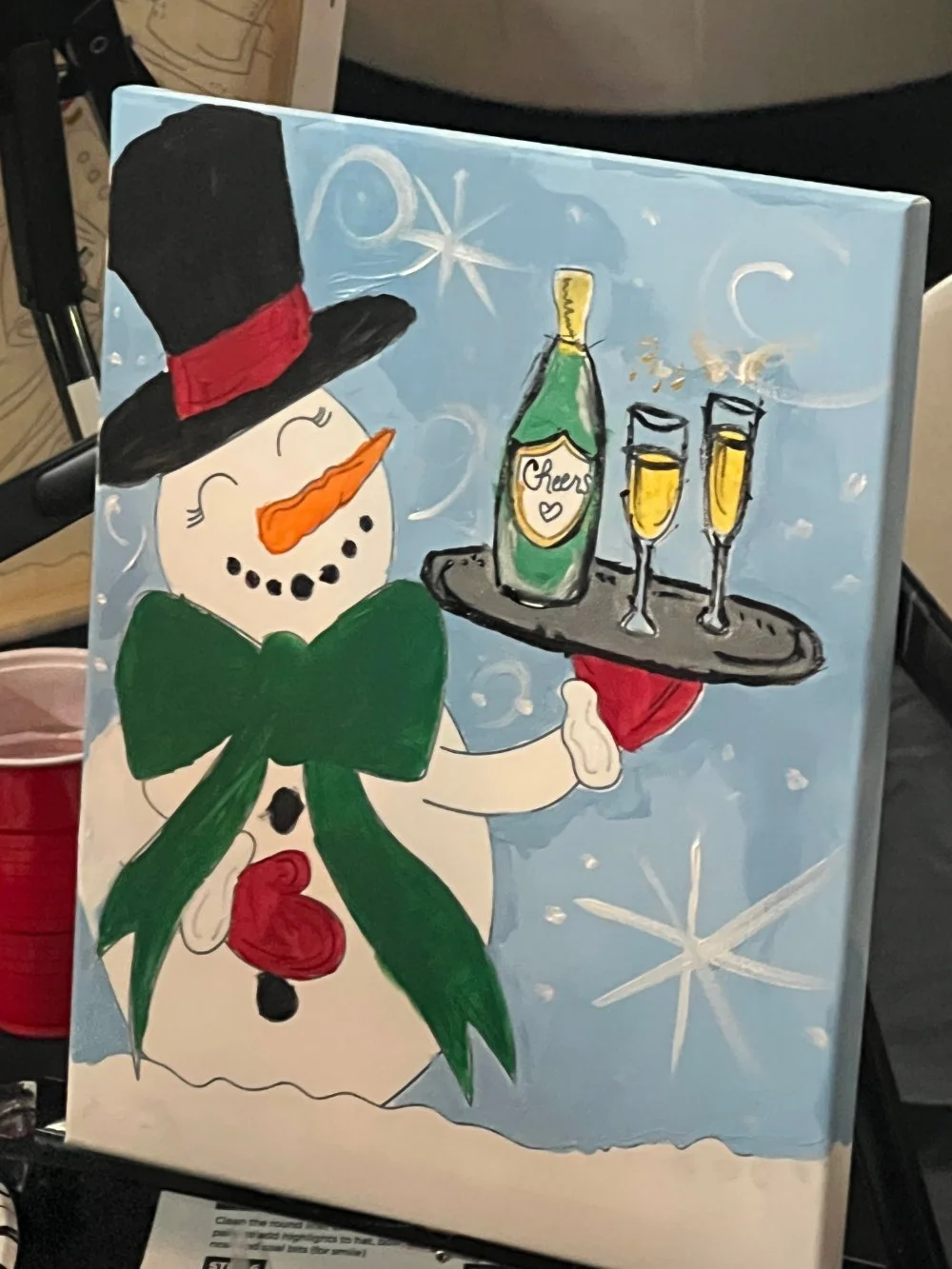 A cheerful snowman wearing a top hat with a red band, a green bow, and buttons, holding a tray with a bottle labeled 'Cheers' and two glasses of champagne, set against a snowy, starry background.