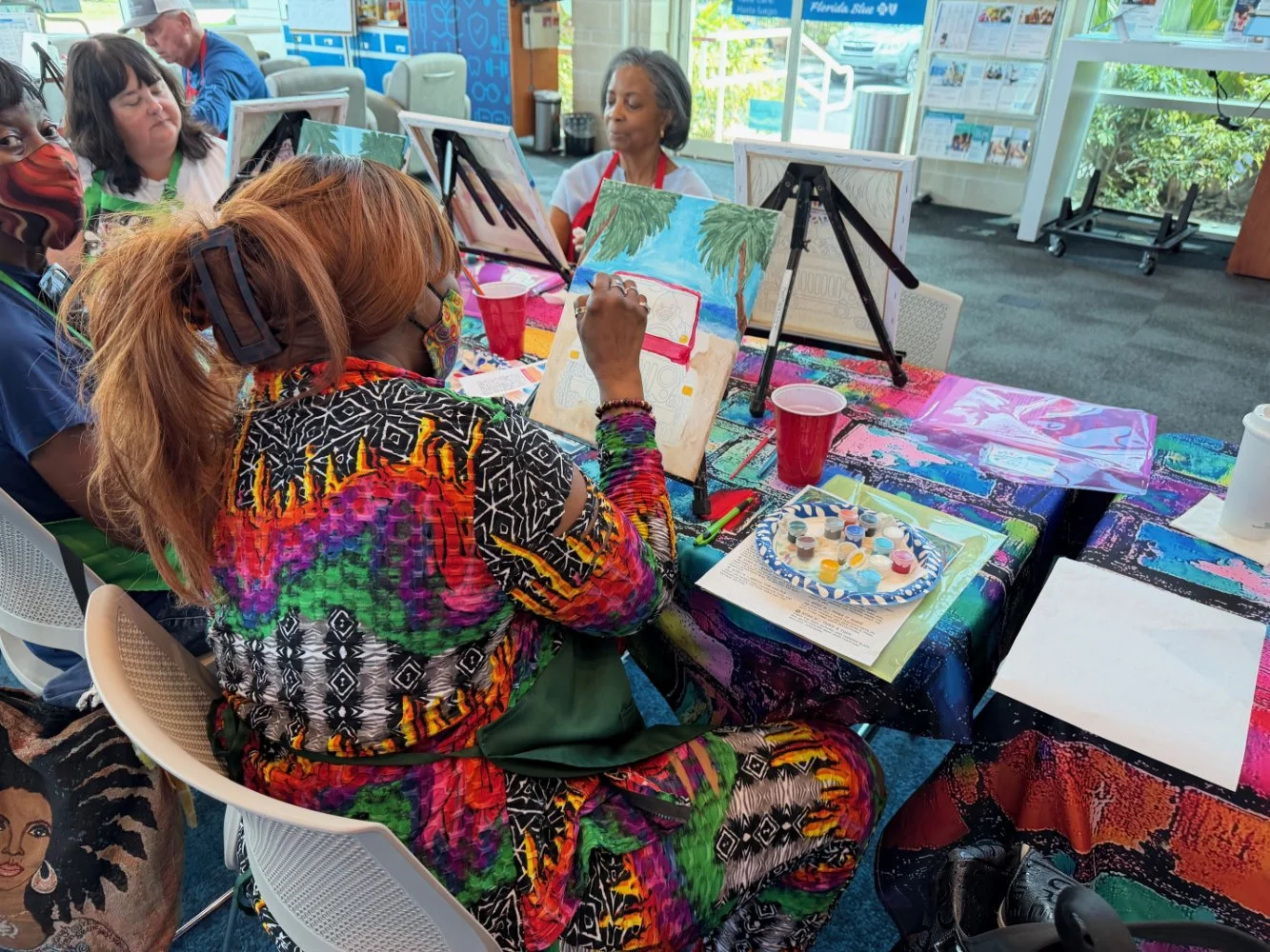 A group of people sitting at a table painting on canvases with a tropical scene, including palm trees and a car, in a well-lit indoor space with large windows and colorful tablecloths.