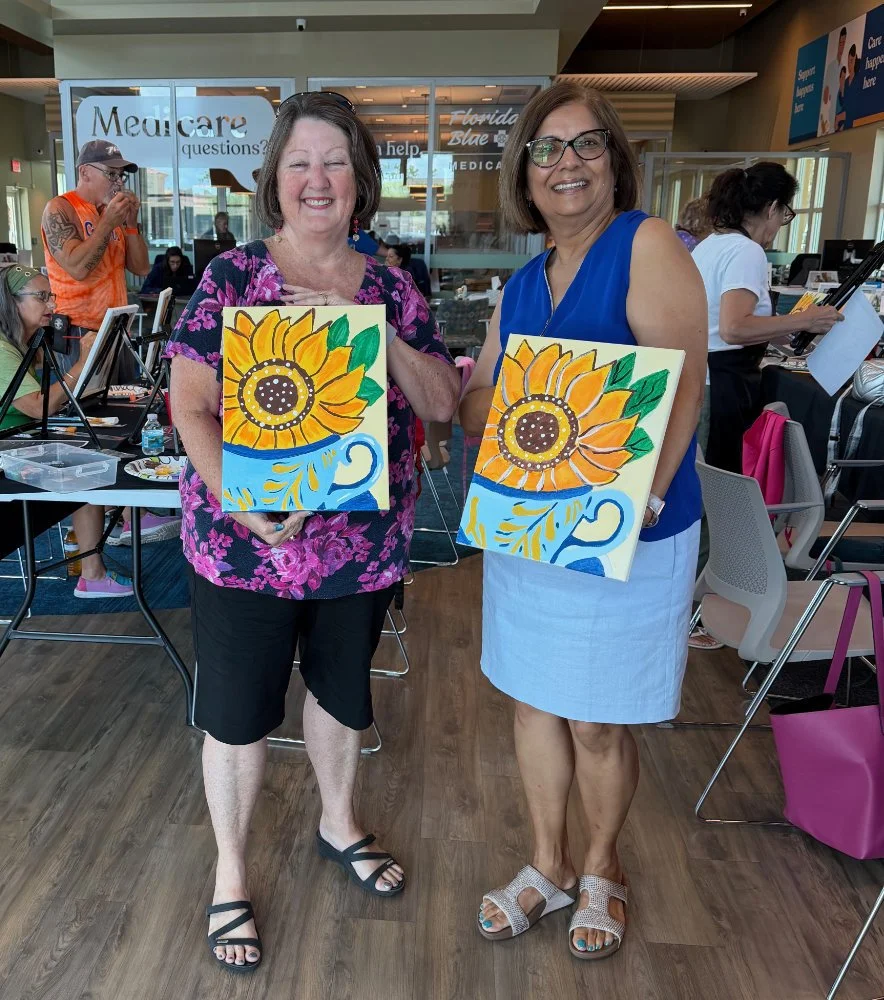 Two women holding paintings of sunflowers in a cafe or workspace. One woman is wearing a floral top and shorts, the other is in a blue blouse and skirt. They are smiling, and other people are working or socializing in the background.
