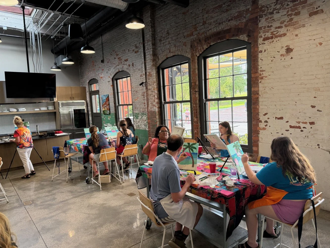 People painting on canvases in a spacious industrial-style room with large windows, exposed brick walls, and colorful tablecloths.