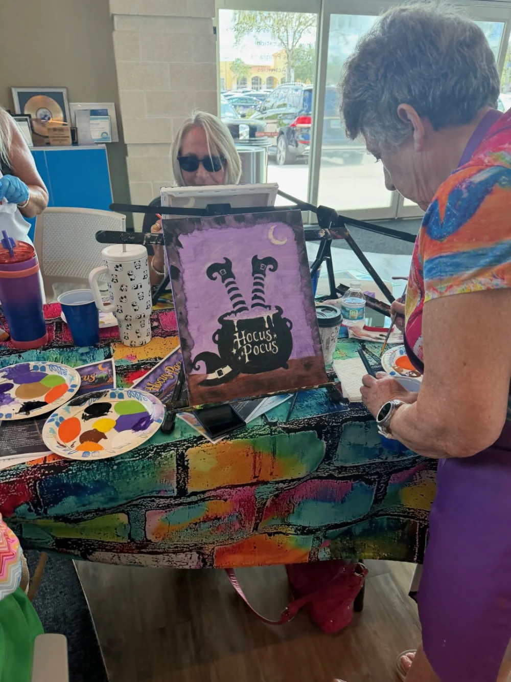 A group of women gathered around a table painting Halloween decorations. One woman is working on a purple canvas featuring a witch's cauldron with striped legs and the words 'Hocus Pocus.' The table is covered with colorful paint palettes, brushes, a