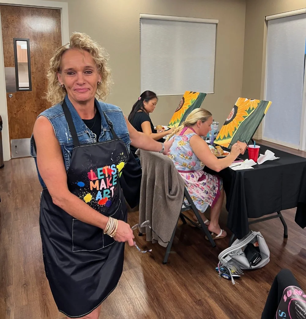 A woman in a denim vest and an apron with colorful paint splashes and the words 'Let's Make Art' stands in a room with her hand on a seated woman painting a sunflower on canvas. Two other women are also painting sunflowers on canvases at a table.