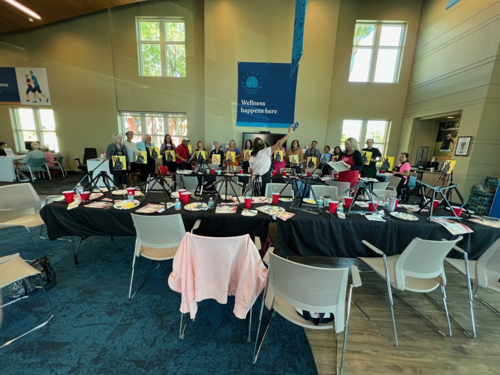 Group of people standing behind a table with photography equipment, including cameras and tripod stands, in a large room with high ceilings, windows, and a blue banner reading 'Wellness happens here.'