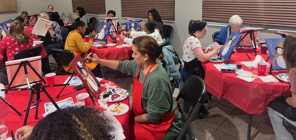 People decorating snowman paintings at a holiday-themed painting class, seated at tables with red tablecloths and painting supplies.