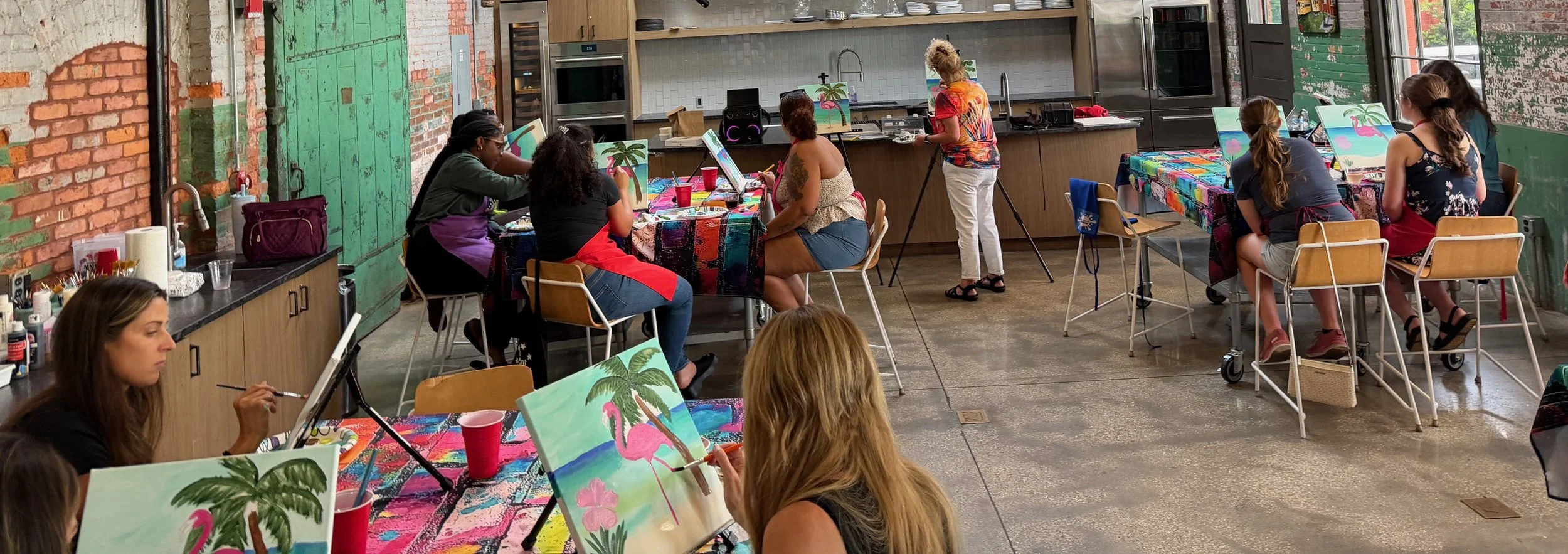 People participating in a group painting class in a spacious industrial-style room with exposed brick walls, some painting tropical scenes with flamingos and palm trees.