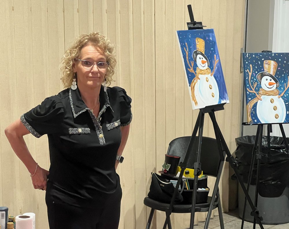 A woman with curly blonde hair, wearing glasses and a black top with embellishments, stands next to two easels with snowman paintings, in an indoor setting with beige wooden panel walls.