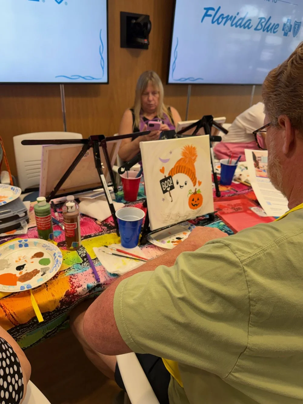 People painting Halloween-themed pictures at a table decorated with a colorful tablecloth, with pens, brushes, and containers of paint around them, at a craft event. A woman in the background is looking at her phone, and there are screens with 