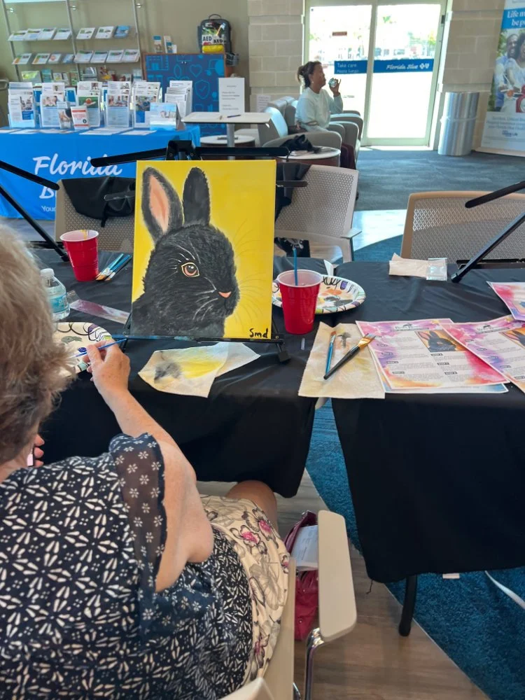 A woman painting a black rabbit on a yellow background at an indoor event space, with art supplies, tables, and informational displays visible.