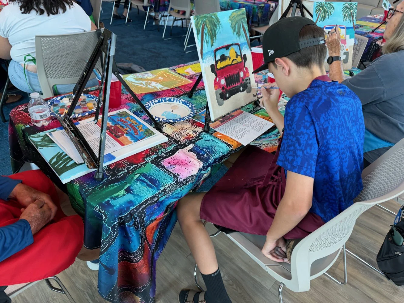 A young boy painting a picture of a red Jeep with palm trees in the background at a painting workshop.
