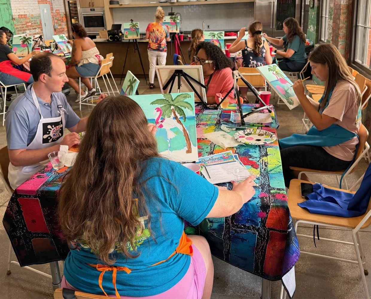 People participating in a paint and sip art class, painting tropical beach scenes with pink flamingos and palm trees on canvases. The room is decorated with a colorful tablecloth and large windows letting in natural light.
