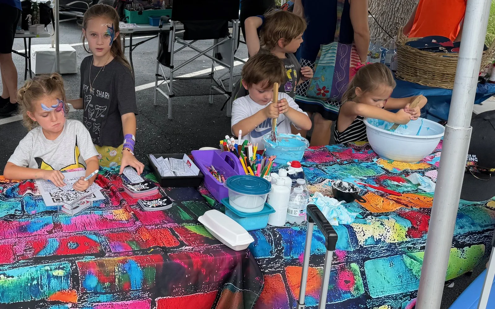 Children at a colorful table engaged in arts and crafts activities, with face paint, markers, and bowls of materials.