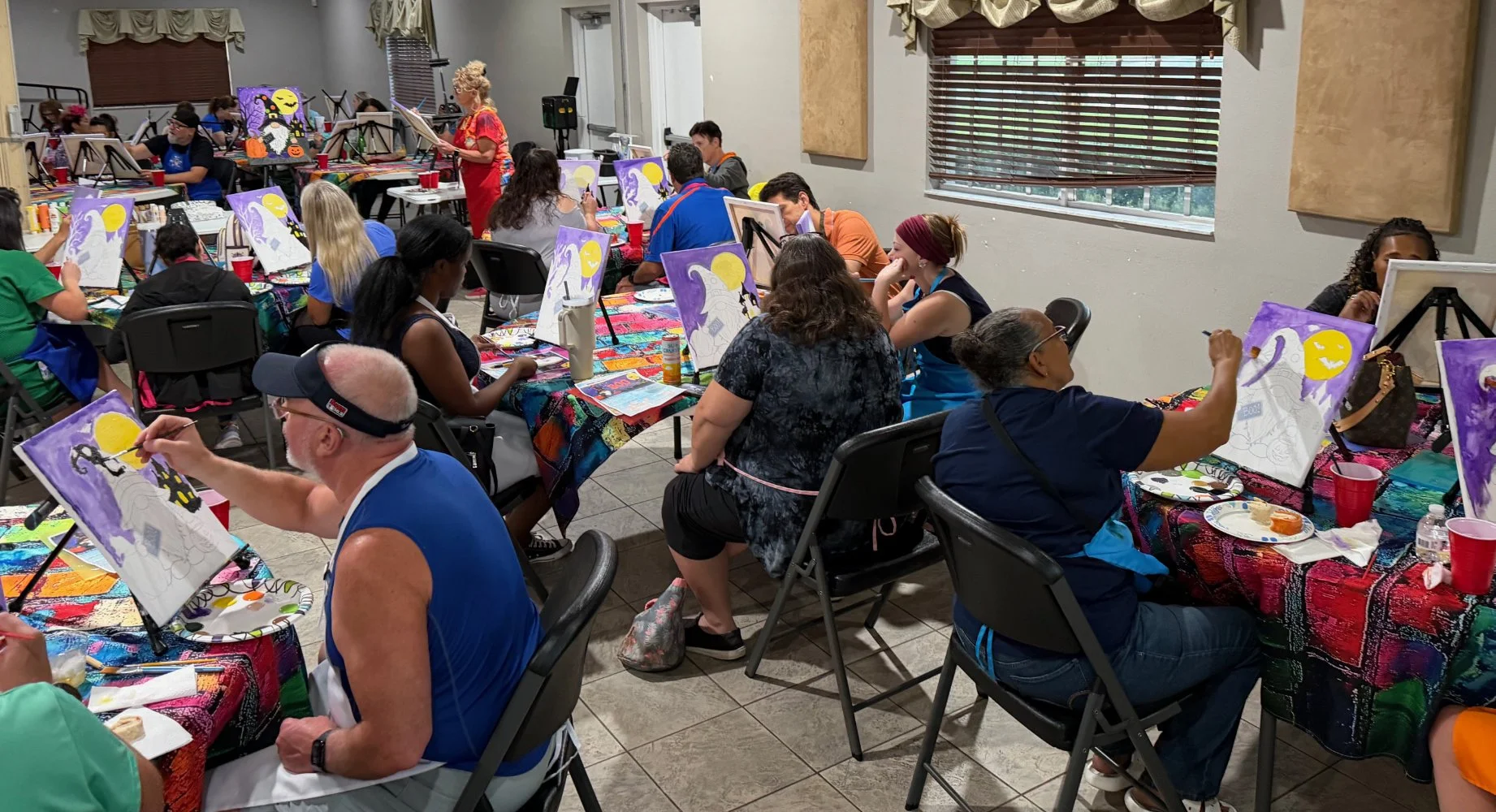 People seated at tables painting Halloween-themed pictures on canvases during a group art activity in a well-lit room with large windows and colorful tablecloths.