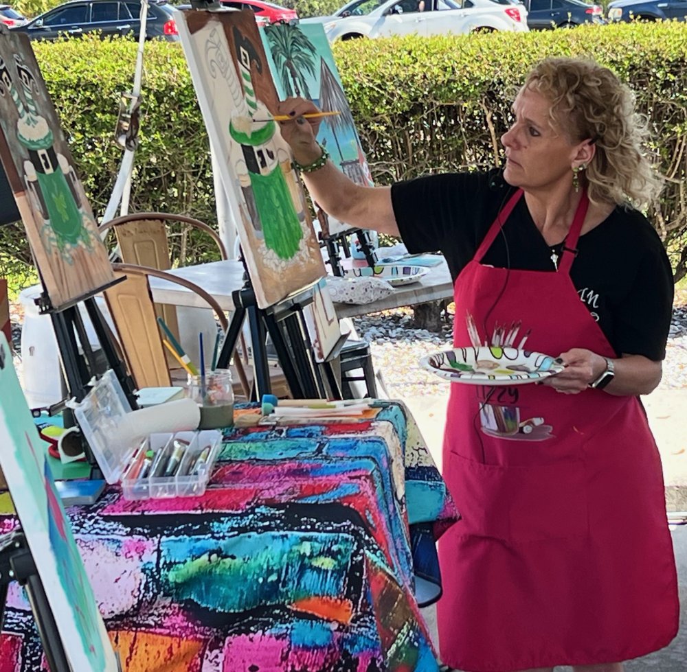 Woman wearing a pink apron painting a tropical scene on a canvas outdoors.