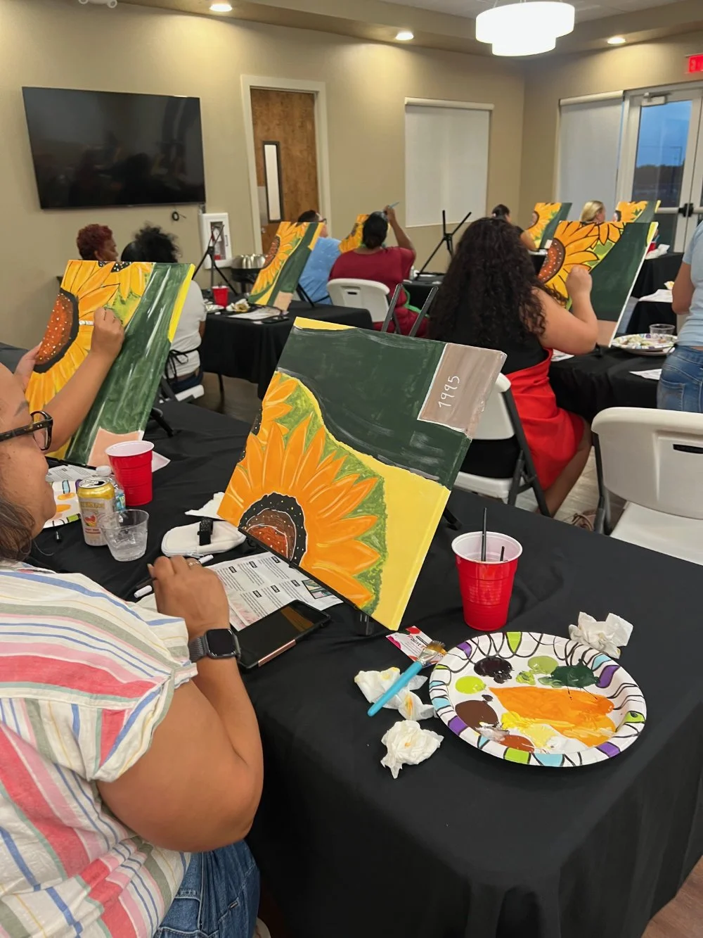 A group of people participating in a painting class, each working on canvas paintings of sunflowers on black tables with painting supplies, in a well-lit indoor room with windows and a large TV on the wall.