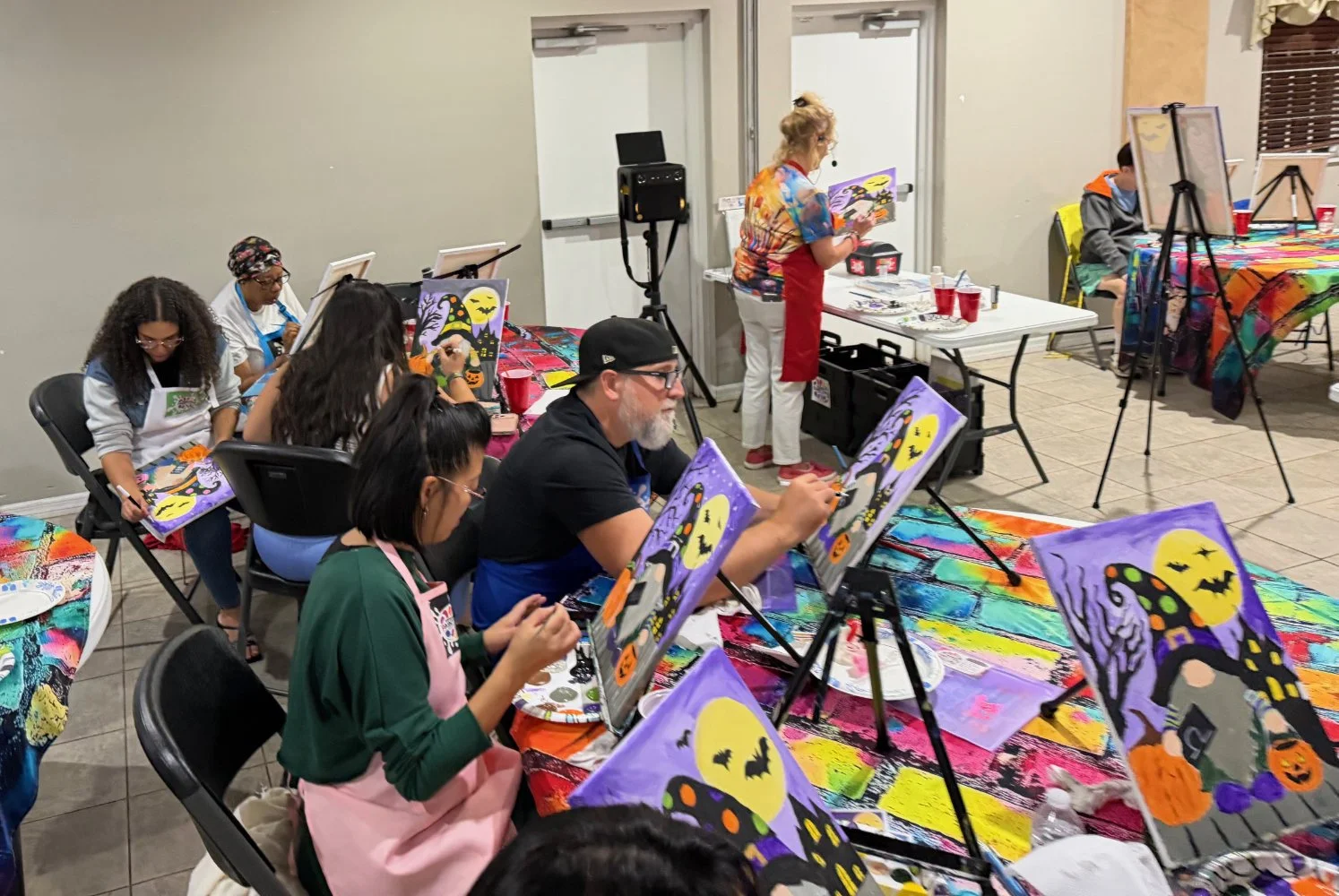 Group of people painting Halloween-themed pictures on canvases at a paint and sip event, with a woman leading the class.