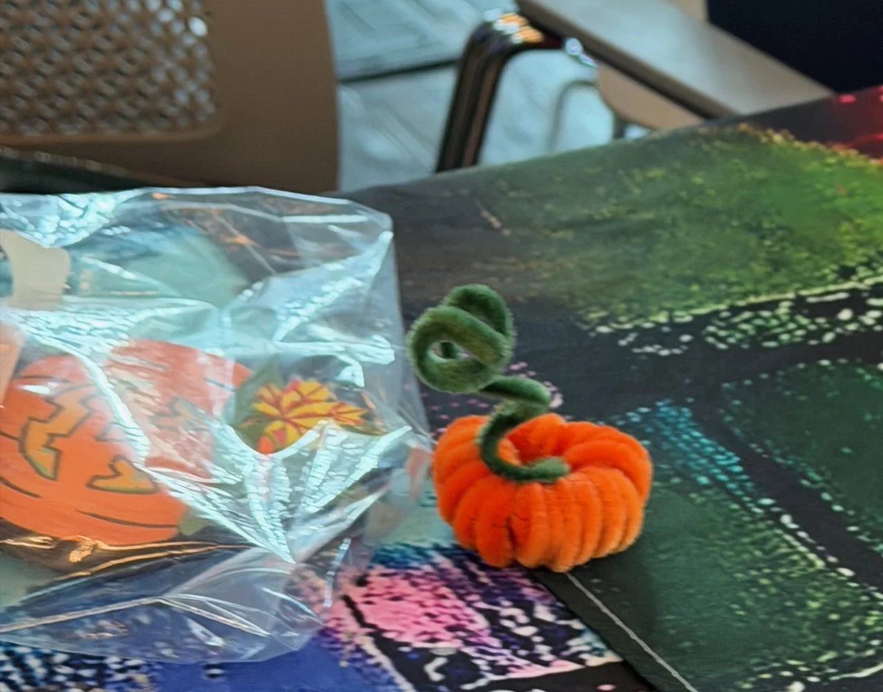 A small plush pumpkin with a green curly stem sits on a colorful tablecloth. A clear plastic bag with orange and yellow pumpkin decorations is nearby.