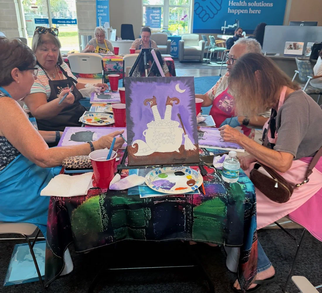 A group of people participating in a painting class, painting a Halloween-themed picture of a cauldron with floating objects and a moon in the background. They are seated around a table covered with a colorful cloth, with art supplies and drinks visi