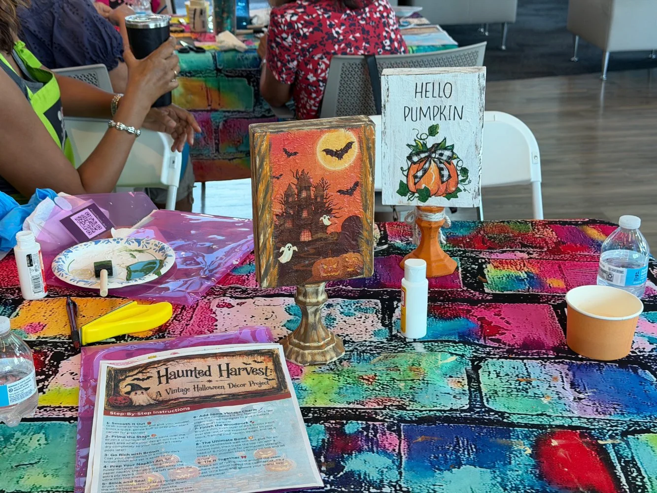 Table with Halloween-themed decorations, including a haunted house and ghost painting, a sign that reads 'Hello Pumpkin' with a pumpkin illustration, craft supplies like glue, scissors, paper cups, water bottles, and an instruction sheet titled 'Haun