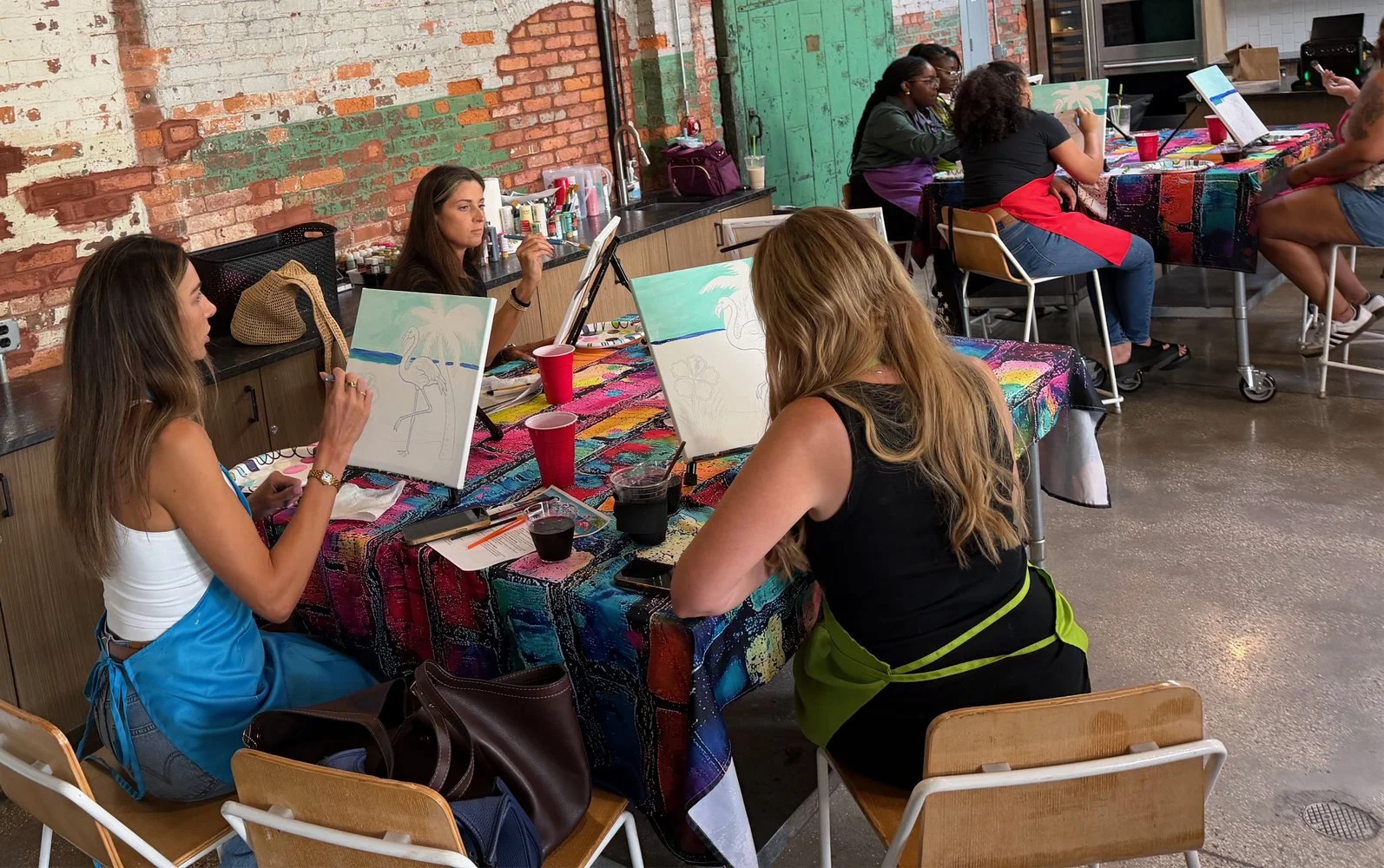 Women participating in a group painting class, sitting at a long table with colorful tablecloths, painting canvases with flamingo and beach scenes, in a studio with brick walls and an industrial style interior.