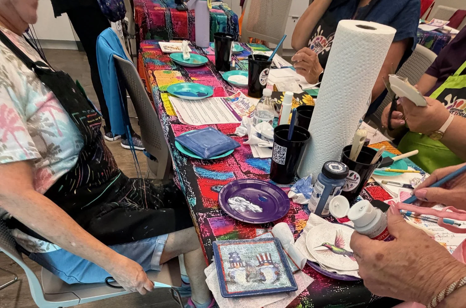 People gathered around a table covered with a colorful tablecloth, engaged in arts and crafts with various supplies including paper towels, paint bottles, scissors, and paper plates.