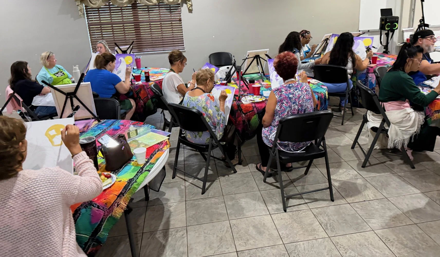 A group of women participating in a painting class in a room with tables covered in colorful tablecloths. They are seated, focusing on their canvases, with some painting and others observing or taking notes. The room has a window with blinds and art 